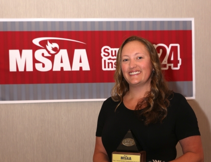 a woman stands in front of a msaa sign, holding an award.