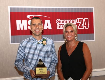 two people stand in front of a msaa sign, one is holding an award.
