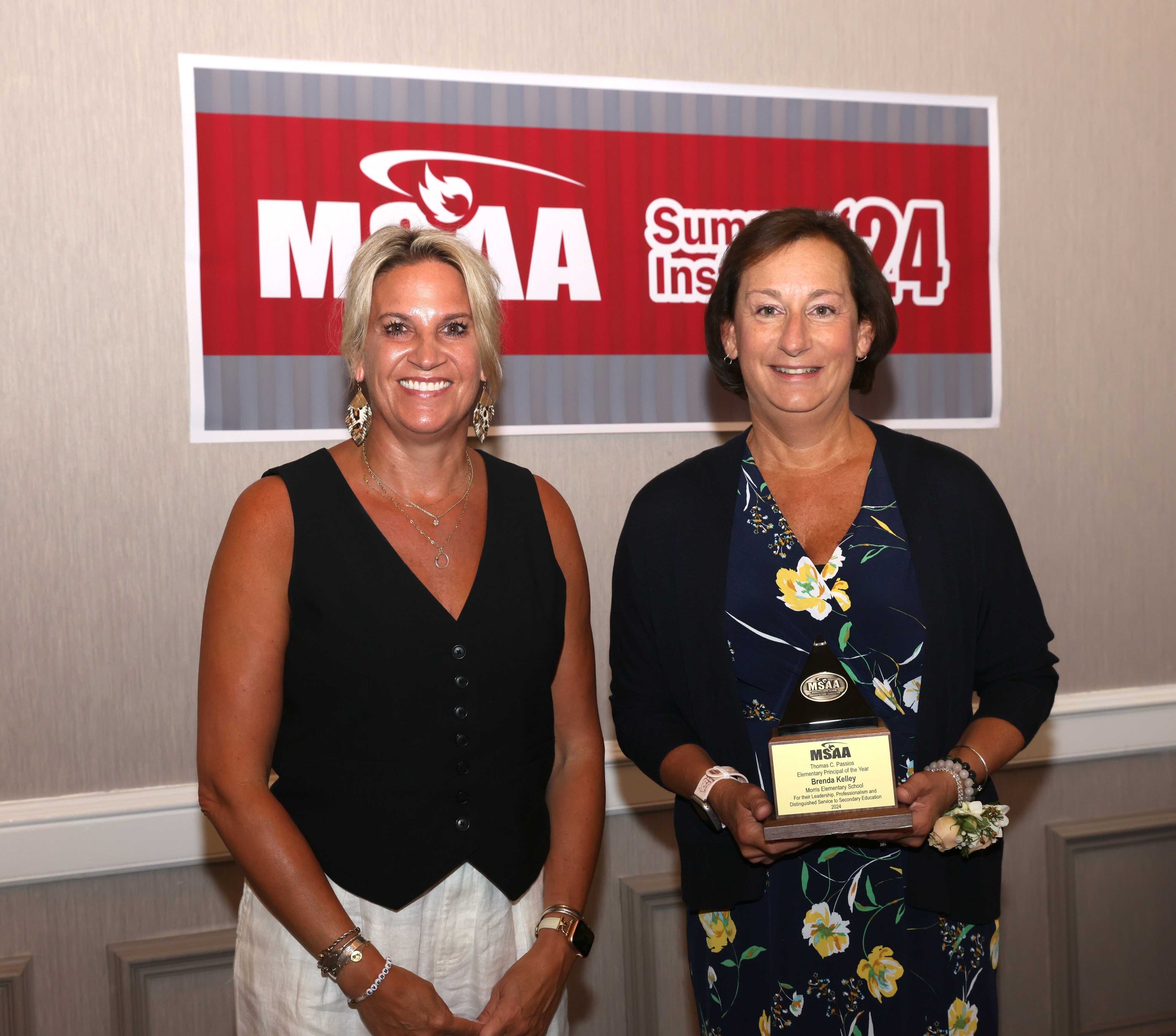 two people stand in front of a msaa sign, one is holding an award.