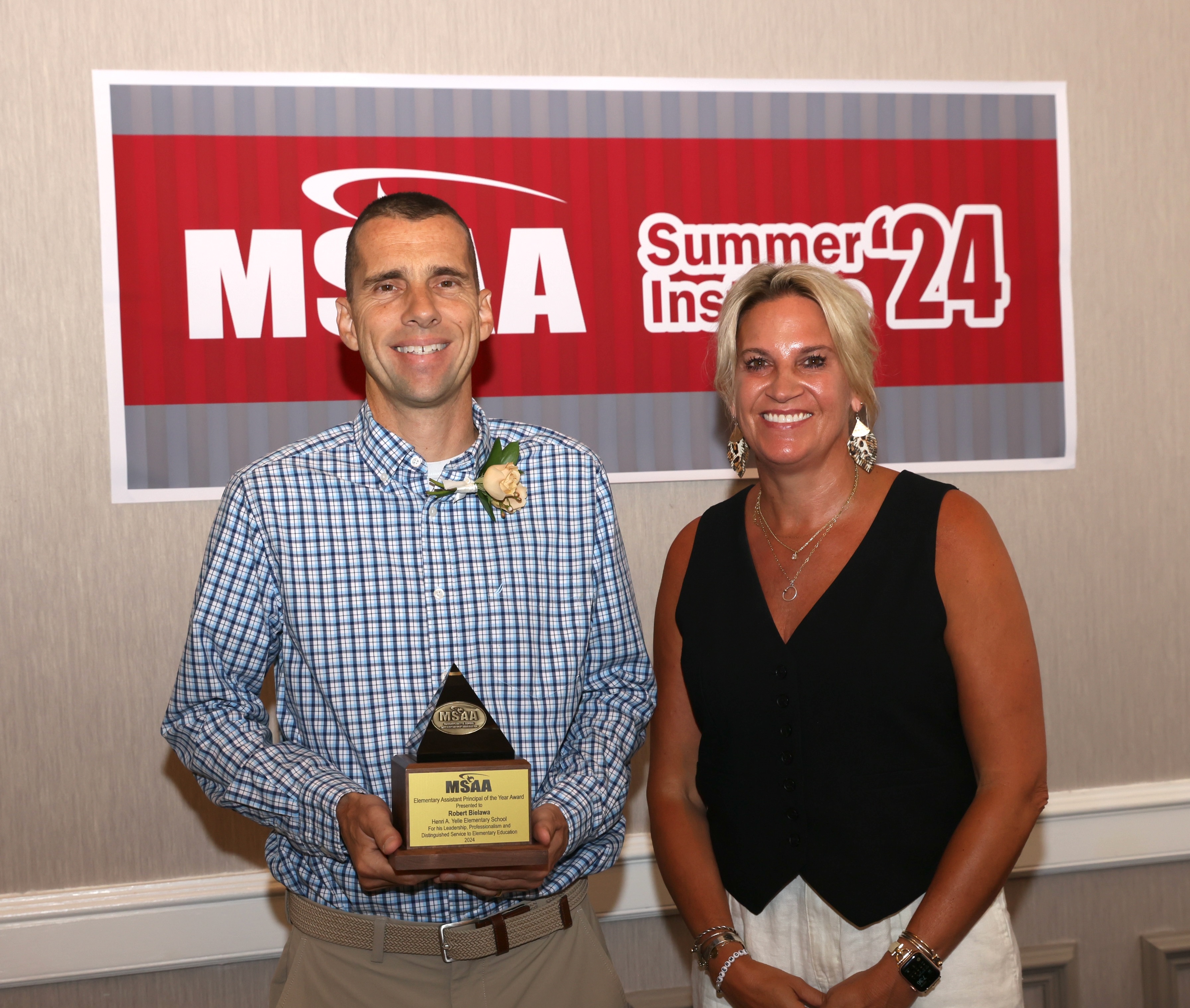 two people stand in front of a msaa sign, one is holding an award.